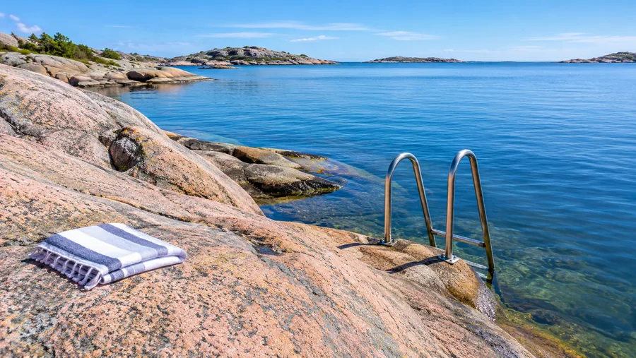 Rocas de granito lisas y rosáceas que descienden hacia un mar azul transparente, con una escalera de metal para bañarse fijada a la roca